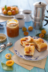 Tavolo imbandito con quadrotti di dolce farcito di  confettura di susine e con sullo sfondo tazze di caffè, caffettiera,  vasetto di confettura e frutta fresca sparsa e su piatto