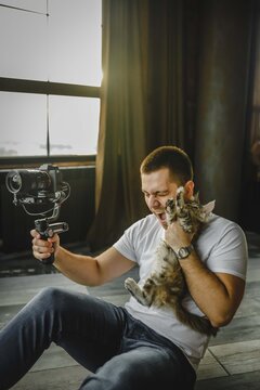 Handsome young european man holds on shoulder grey aine coon cat. guy in white T-shirt holding camera with electronic stabilization and holding cat