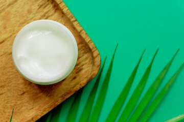 White jar of face cream on a wooden tray. green background, top view, tropical leaves. Concept of natural cosmetics and spa.