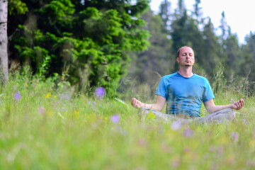 Obraz premium Man is meditating on green grass in the nature with face raised up to sky and eyes closed on sunny summer day. Concept of meditation, dreaming, wellbeing and healthy lifestyle.