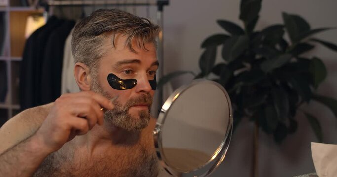 Mature guy drinking alcohol before plucking eyebrows in front of mirror. Handsome bachelor with eye patches making painful facial expression while doing beauty procedure at home.