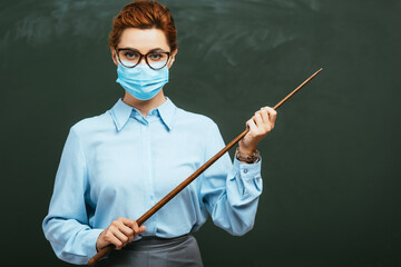 young teacher in medical mask looking at camera while holding pointing stick near blank chalkboard