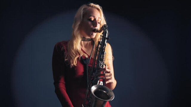 Young beautiful woman in red concert dress playing a melody on saxophone over dark background in studio