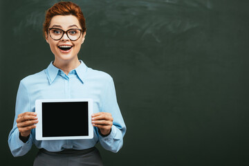 excited teacher showing digital tablet with blank screen near chalkboard