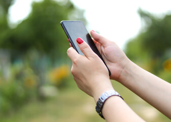 Mobile phone in a female hand with red manicure. Blurred green bokeh background.