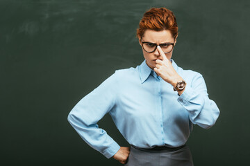 serious teacher touching eyeglasses while standing with hand on hip near chalkboard