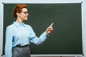 smiling teacher looking away and pointing with finger near chalkboard