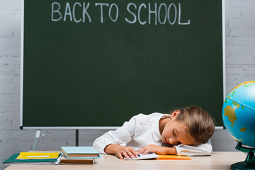 exhausted schoolgirl sleeping at desk near globe and chalkboard with back to school lettering