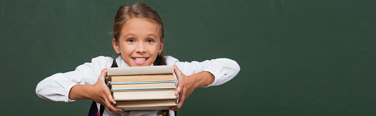 horizontal image of happy schoolgirl showing stack of books near green chalkboard