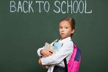 discouraged schoolgirl with backpack holding books near chalkboard with back to school inscription