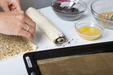 A woman rolls up puff pastry with walnuts into a tube. For making puff pastry curls with poppy and walnut filling. Nearby on the table are ingredients and tools.