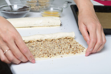 A woman rolls up puff pastry with walnuts into a tube. For making puff pastry curls with poppy and walnut filling. Nearby on the table are ingredients and tools.