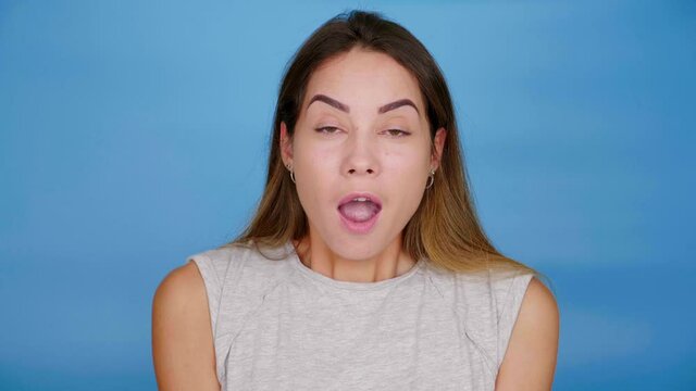 Beautiful woman in gray T-shirt shocked looking camera on blue background with copy space. Girl surprised with mouth open. 4K slow motion footage