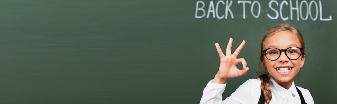 horizontal image of cheerful schoolgirl in eyeglasses showing thumb up near chalkboard with back to school inscription