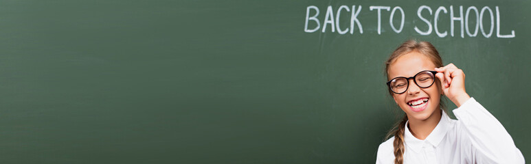 panoramic crop of excited schoolgirl laughing with closed eyes and touching eyeglasses near chalkboard with back to school lettering - obrazy, fototapety, plakaty