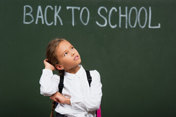 selective focus of pensive schoolgirl touching head while looking up near chalkboard with back to school inscription