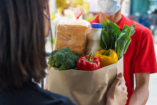 Woman Customer Receiving Fresh Food Set Bag From Food Delivery Service Man With Protection Face Mask In Red Uniform At Door Home, Express Delivery, Quarantine, Virus Outbreak, Food Delivery Concept