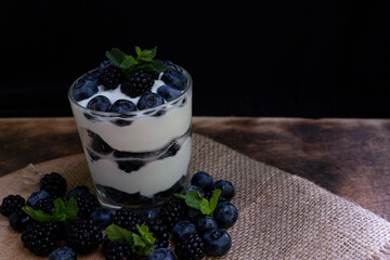 Cottage cheese dessert with blueberries, blackberries and mint in a transparent glass glass. On a burlap napkin and on a wooden background. Delishious.