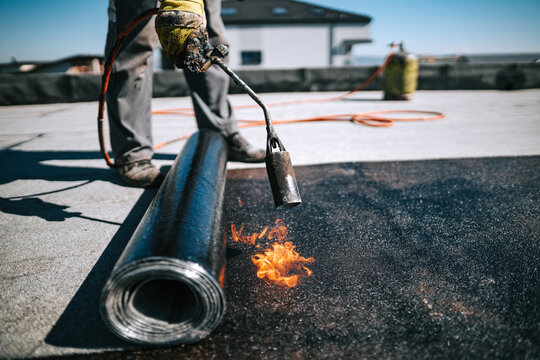 Professional workers insulating rooftop with bitumen membrane. Waterproofing details at construction site. Bituminous membrane waterproofing system details and installation on flat rooftop.