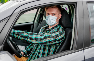 A young man in a protective sterile medical mask is sitting behind the wheel of the car. COVID-19.