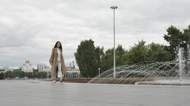 A girl with long hair and a brown trench walks along the fountain on a summer warm evening. High quality 4k footage