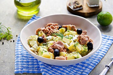 Potato salad in a Mediterranean style with canned tuna and olives in a ceramic bowl on a light gray concrete background. American cuisine.