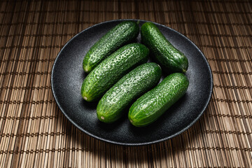 fresh green vegetables cucumbers lie in a ceramic black textured plate on a bamboo path
