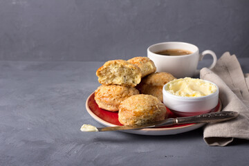 Homemade freshly baked scones with cheese and herbs on a red plate with butter, a cup of coffee on a gray textured background