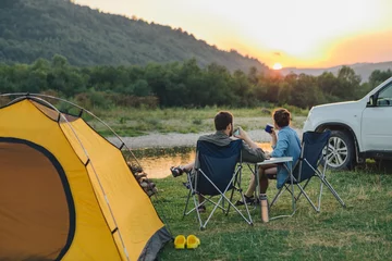 Papier Peint photo Lavable Camping couple sitting in camp chairs looking at sunset above river in mountains