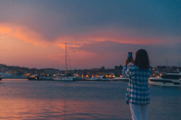 Fototapeta premium woman taking picture of the sunset over sea dock on her phone