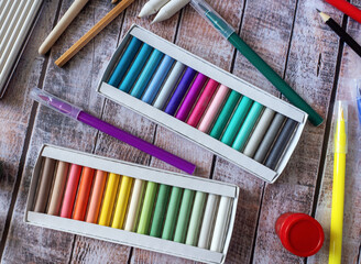 The boxes contain a set of pastels. Felt pens, pencils and other items lie on a wooden table....