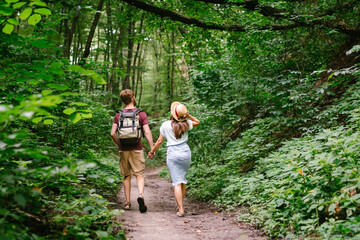 Happy caucasian couple are walking in a dense forest along the path holding hands, rear view. Hikers with backpack looking for place for picnic in wooded area in the summer. Walk outdoors together