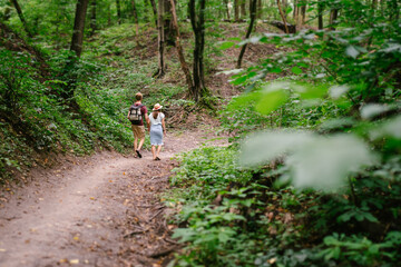 Happy caucasian couple are walking in a dense forest along the path holding hands, rear view. Hikers with backpack looking for place for picnic in wooded area in the summer. Walk outdoors together