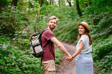 Guy and girl walk together in wood along the trail, holding hands. Back view. Happy couple holding hands and walking forest path. Hikers walk in wooded area with a backpack in summer