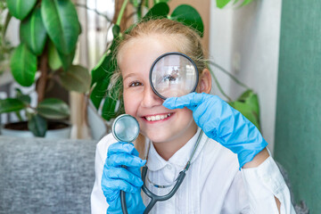 A Caucasian girl of 9-10 years old in a white coat, blue medical gloves holds a stethoscope in her hand, looks through a magnifying glass, smiles. Research work, medicine, health. Medical clinic.