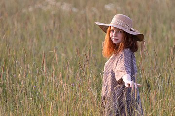 Obraz premium emotional redhead girl playing in the grass on a summer meadow at sunset in an elegant dress
