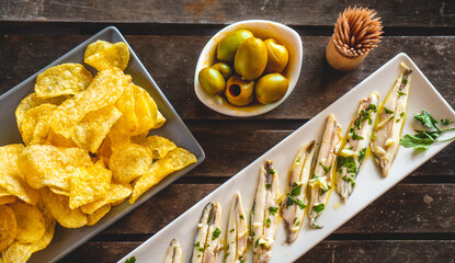 Three dishes with potato chips, picked anchovies and green olives on a dark wooden table with toothpicks. Typical Spanish snacks to eat in a bar. 