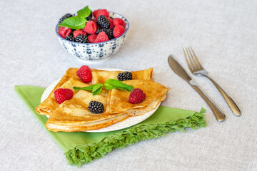 Delicious sweet crepes decorated with raspberries, blackberries and mint leaves on a white plate, next to a knife, fork and green napkin