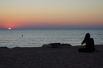 Naklejka na ściany i meble Silhouette of a man sitting on a deserted beach and photographing the sea during sunset