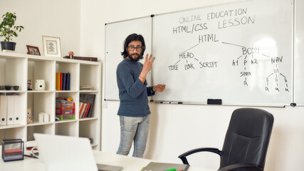 Obraz na płótnie Canvas Teaching code programming. Young bearded male teacher pointing at whiteboard and explaining how to code HTML CSS, giving lesson online at home