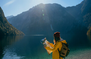 a girl in a yellow jacket and a red hat with a backpack in sunny weather looks at a map in the mountains near a blue turquoise lake side view