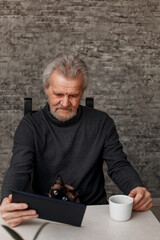 An elderly man drinks coffee in the kitchen and uses a digital tablet
