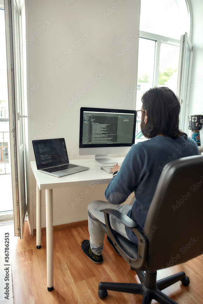 Wall mural rear view of young man, software developer writing code on desktop computer while sitting at his wor