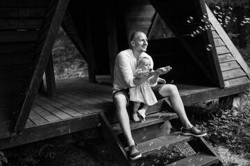 Caring father hold little daughter sitting on stairs, cute toddler with dad try to catch the bubbles, happy family moments, childhood concept
