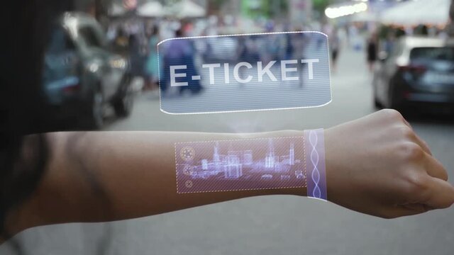 Female hand activates conceptual HUD holograms on smart bracelet with text E-ticket. Unrecognizable woman with holographic technology on a city street background