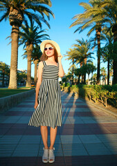 Attractive woman wearing a striped dress and summer straw hat outdoors over palm trees background