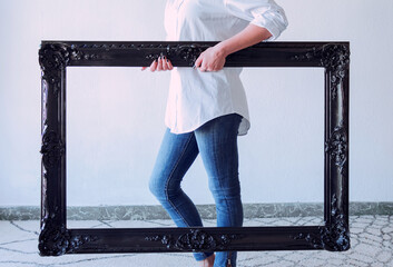 Plano frontal de una mujer con camisa blanca y jeans azules posando junto a una pared blanca como fondo y sosteniendo con sus manos un marco vintage y negro de una antigua pintura