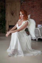 Pretty, slender woman with wings. Female in stage image of an angel. Emotions of girl in dress sitting on chair in an old textured interior room. Actress shows her abilities. Author space in photo