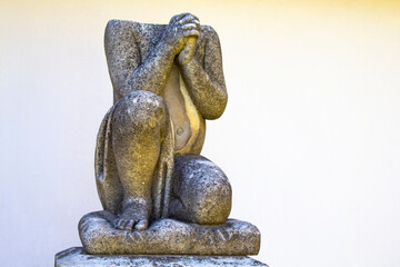 Decapitated statue of child kneeling angel with arms crossed against blue sky and cemetery...