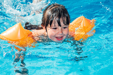 Little black-haired girl smiling and swimming in the pool wearing sleeves
